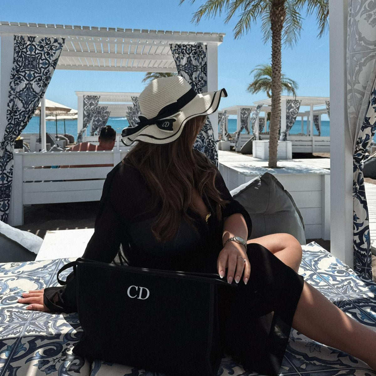 Woman sitting on a sunbed with a black bag featuring a personalised hat, in a beachside setting.