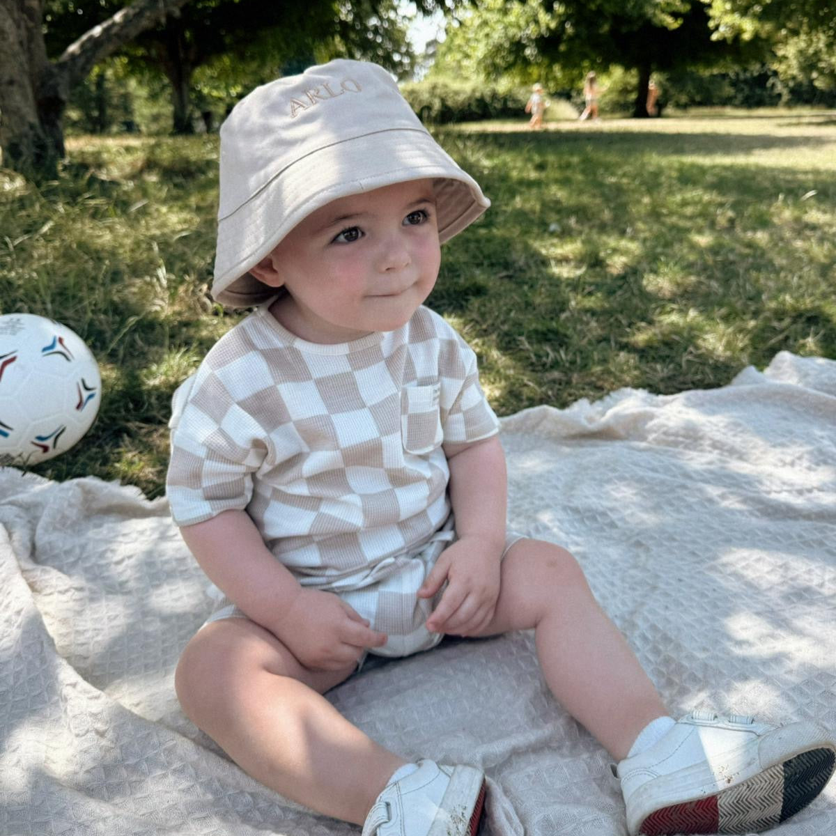 Baby sitting on a blanket outdoors wearing a checkered outfit and beige bucket hat.