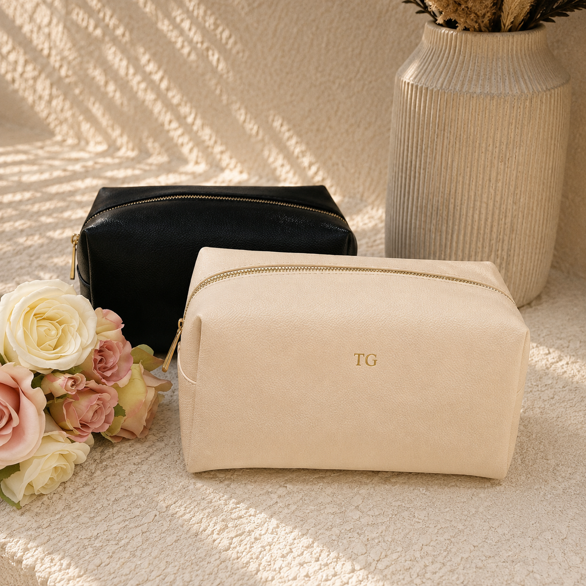 Two zippered cosmetic pouches displayed on a textured beige surface with soft floral props and a ribbed vase in the background. The front pouch is a warm cream/beige color with a gold zipper and a small gold 'TG' monogram, while the rear pouch is black with a gold zipper.