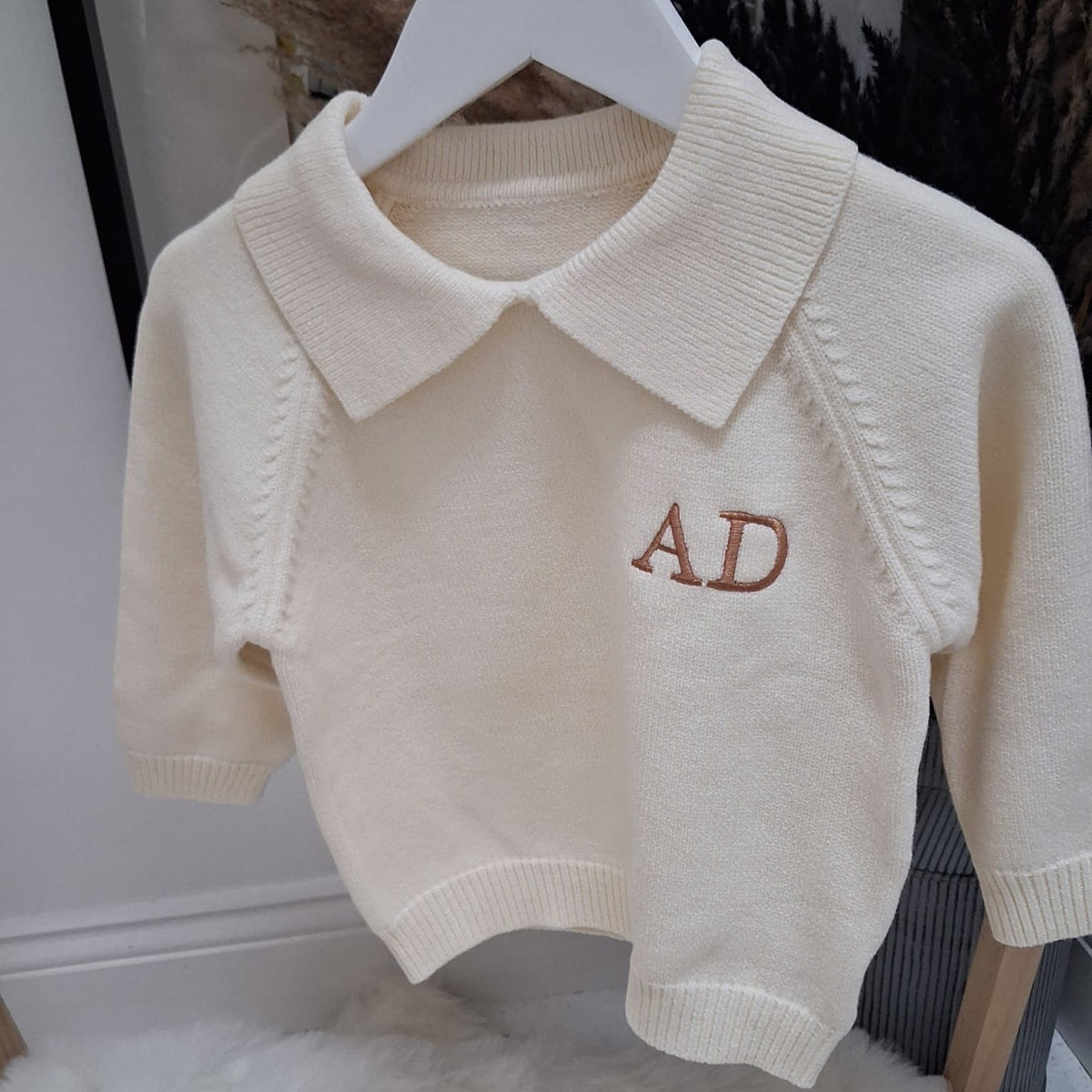 Beige knitted vest with 'AD' embroidery on a white hanger.