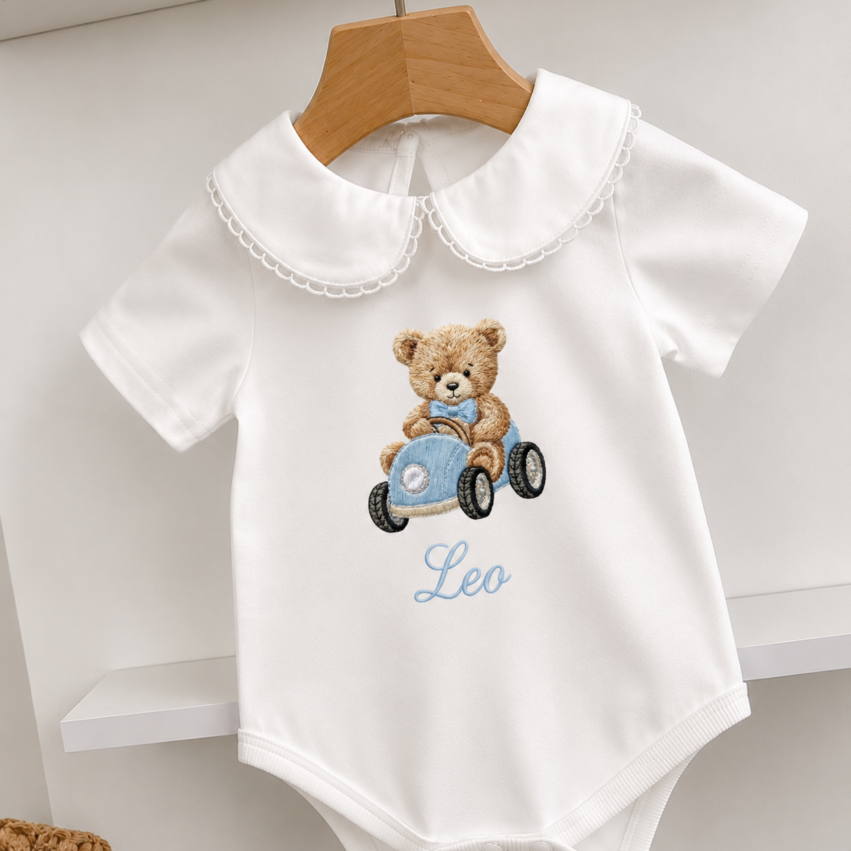A white short-sleeve baby bodysuit hanging on a wooden hanger, featuring a teddy bear driving a blue race car graphic and the name 'Leo' embroidered in light blue script on the front.