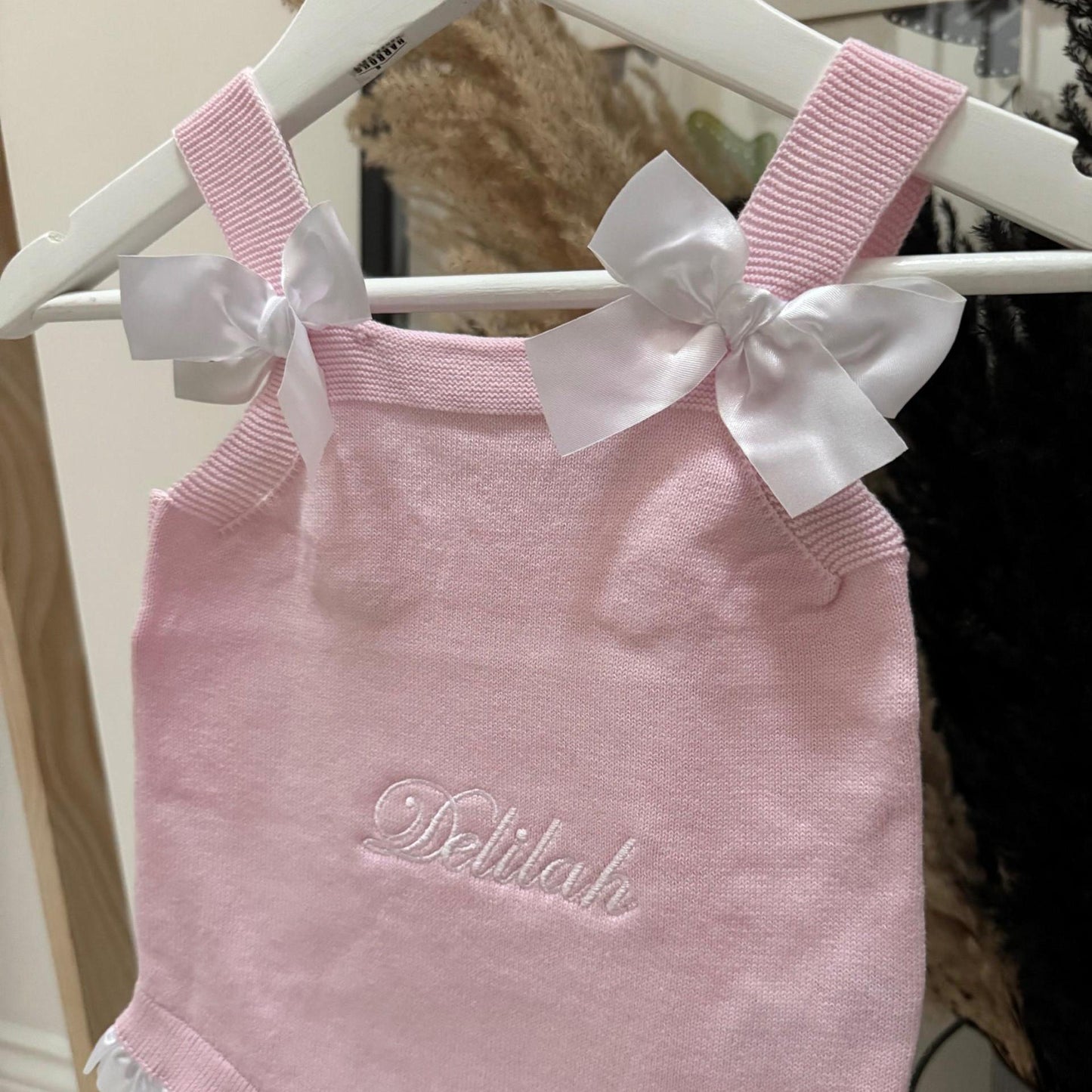 Pink baby romper with white ribbons on a hanger with name Delilah