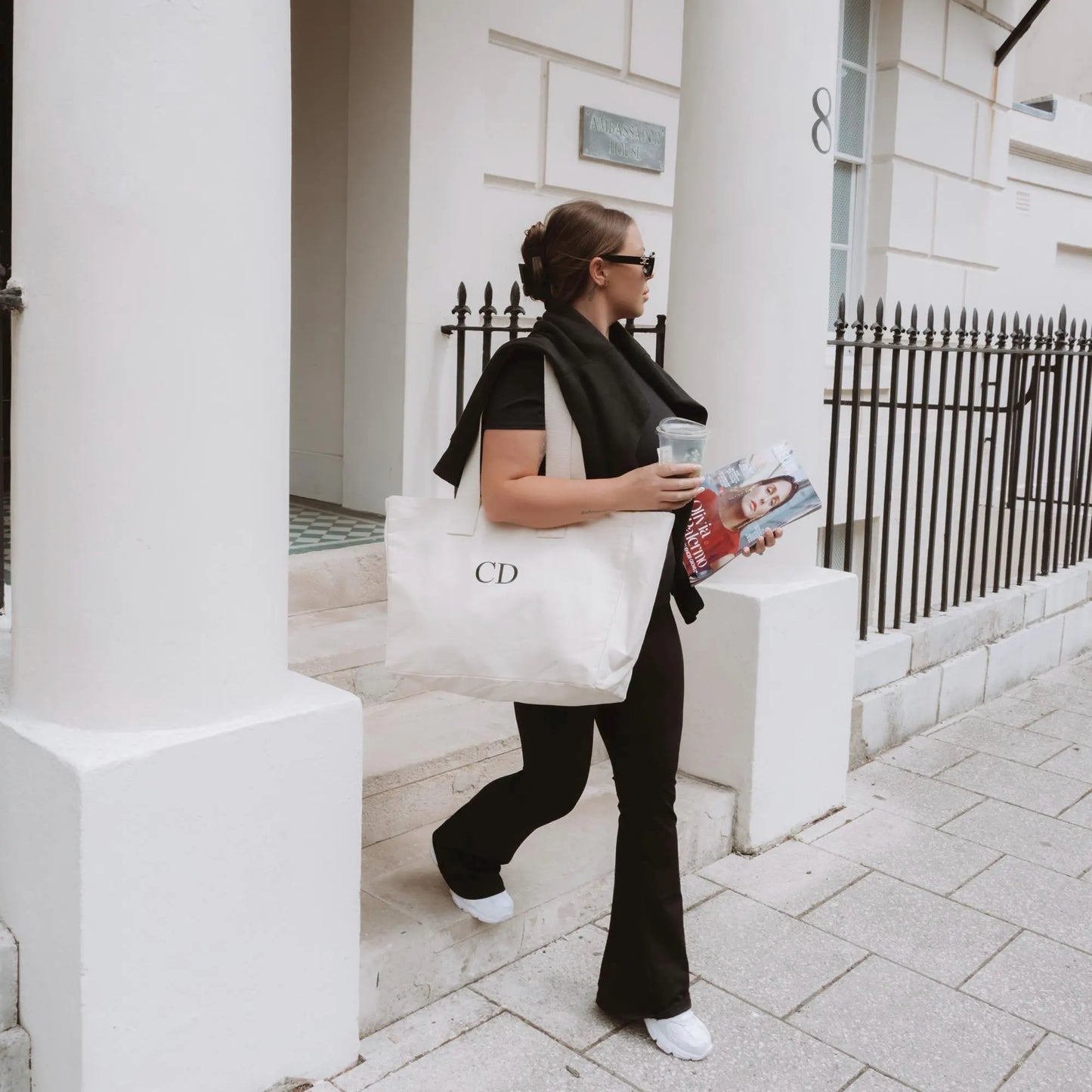 Woman holding a coffee cup and magazine, walking on a sidewalk with a white building in the background