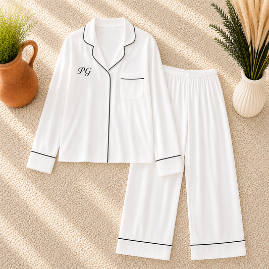 White pajama set with black piping trim, featuring a long-sleeve top with a chest pocket and embroidered initials 'PG' on the left chest, paired with matching wide-leg pants, arranged on a textured beige rug with decorative plants.