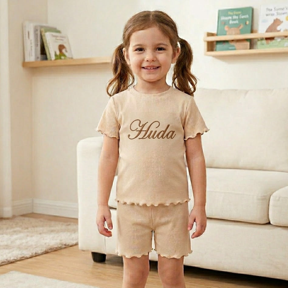 Young girl wearing a beige outfit with 'Huda' printed on it, standing in a living room.