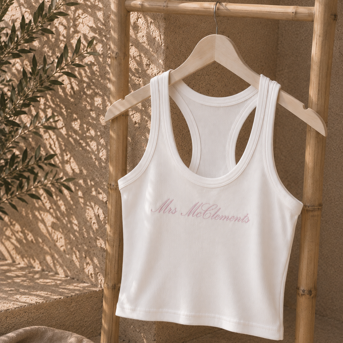 A white racerback tank top with 'Mrs. McElementi' printed in pink cursive across the chest, hanging on a wooden hanger from a bamboo stand in a warm, sunlit rustic setting.