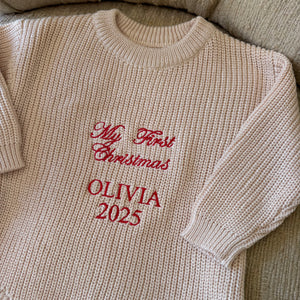Beige baby sweater with red text 'My First Christmas OLIVIA 2025' on a beige surface.