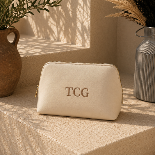 A cream-colored faux leather cosmetic pouch with gold zippers and the letters 'TCG' embossed on the front, styled on a textured stone ledge with potted plants in the background.
