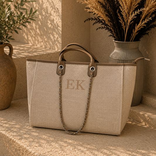 A beige woven tote bag with brown leather handles and a silver chain strap, featuring the monogram 'EK' on the front. The bag is styled on a stone ledge with potted plants and dried grass in the background.