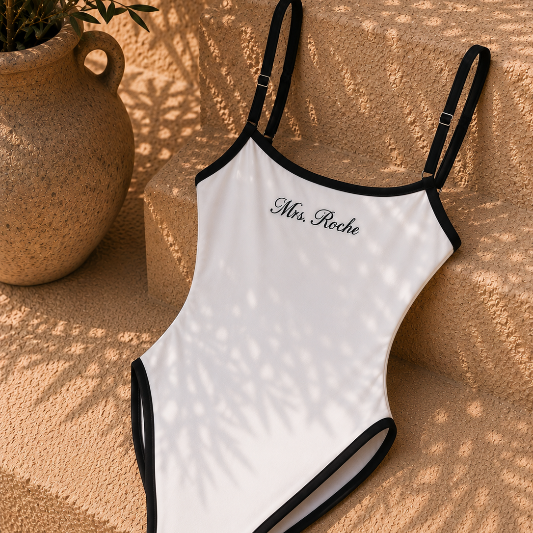 A white one-piece swimsuit with black trim and thin shoulder straps, featuring the text 'Mrs. Roche' printed in black script on the chest.