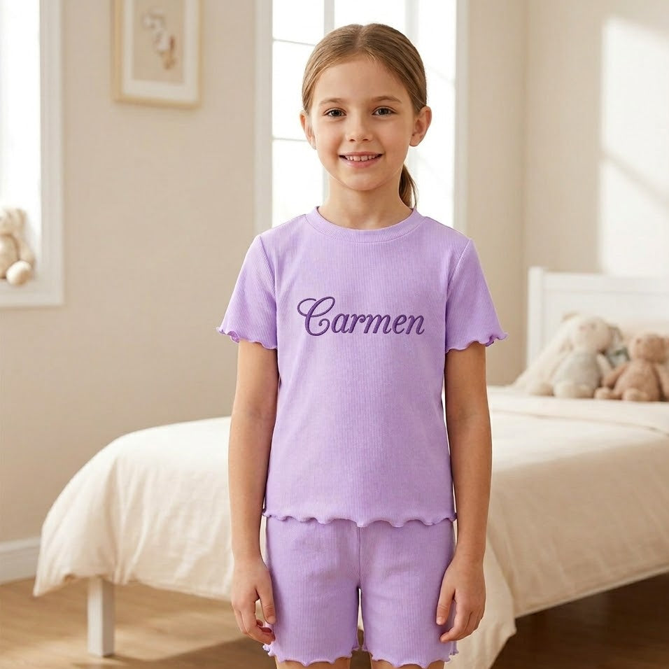Young girl wearing a purple shorts set with 'Carmen' printed on it in a bedroom.