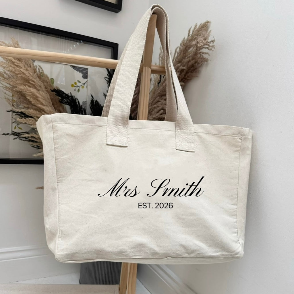 Beige tote bag with 'Mrs. Smith EST. 2026' text against a white wall.