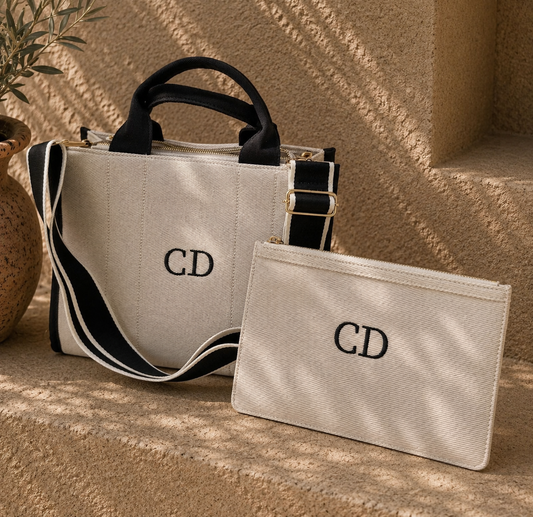 A beige canvas tote bag and matching zip pouch, both embroidered with a black 'CD' monogram, displayed on a textured beige stone surface with dappled sunlight and shadows.