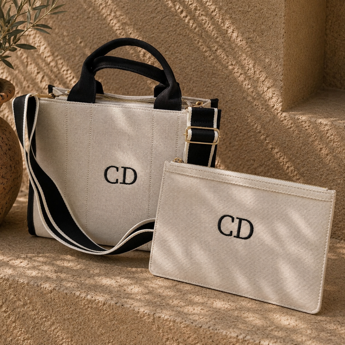 A beige canvas tote bag and matching zip pouch, both embroidered with a black 'CD' monogram, displayed on a textured beige stone surface with dappled sunlight and shadows.