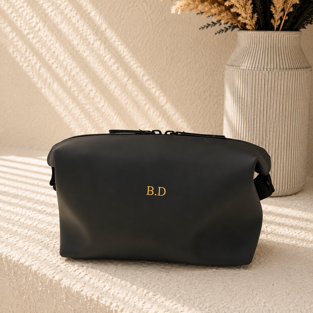 A black faux-leather cosmetic pouch with gold 'B.D' monogram lettering, resting on a textured cream surface with diagonal shadows, next to a ribbed ceramic vase.