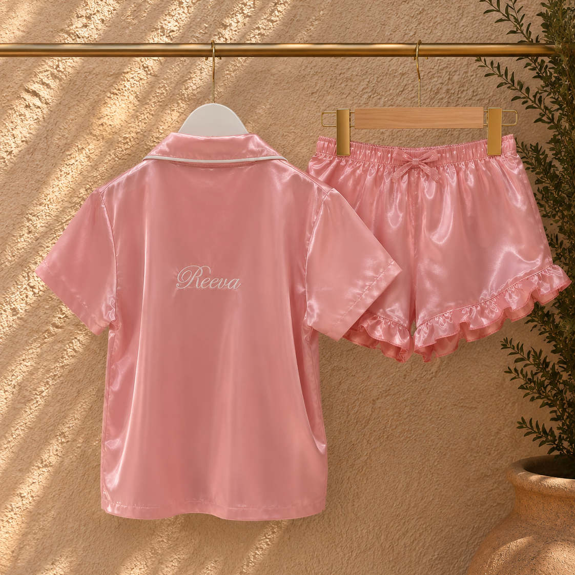 A pink satin short-sleeve collared top and matching pink satin ruffled shorts set hanging on hangers against a textured beige wall with warm sunlight shadows. The top has the word 'Picena' embroidered on the chest.