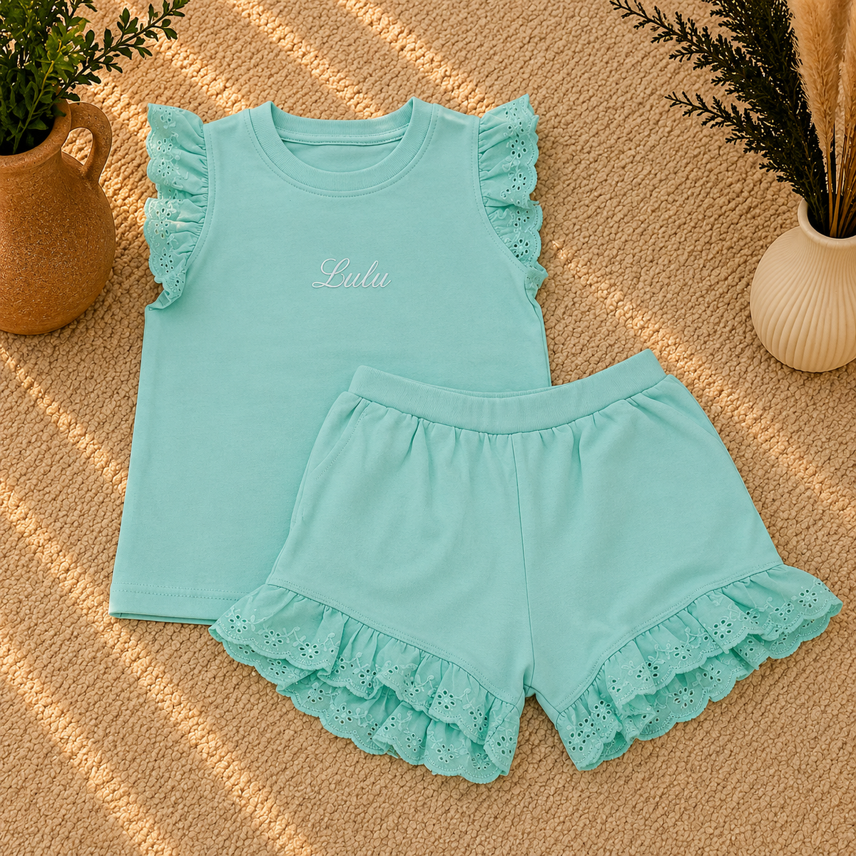 A two-piece children's outfit set in mint green, featuring a sleeveless top with ruffle trim and matching shorts with a ruffled eyelet hem, laid flat on a textured beige surface with decorative plants in vases.