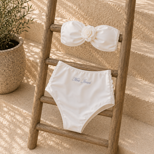 A two-piece womans swimwear set in off-white, styled on a rustic wooden ladder. The top is a bandeau-style bikini top with a large fabric bow and a white fabric flower at the center.