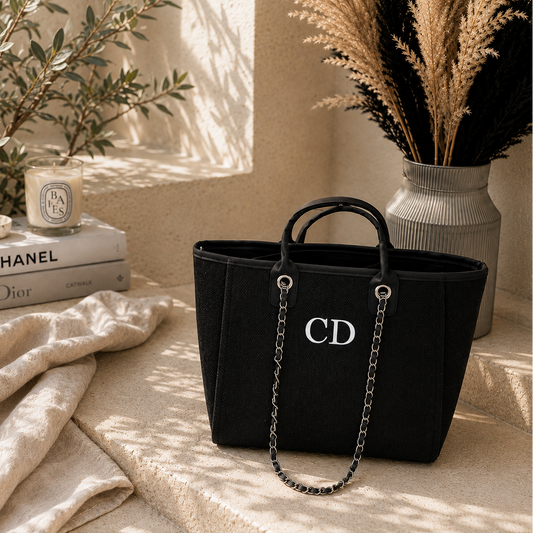 A black canvas tote bag with silver-tone chain handles and a prominent silver 'CD' logo on the front, styled on a beige stone ledge next to a candle, books, and dried grass in a vase.