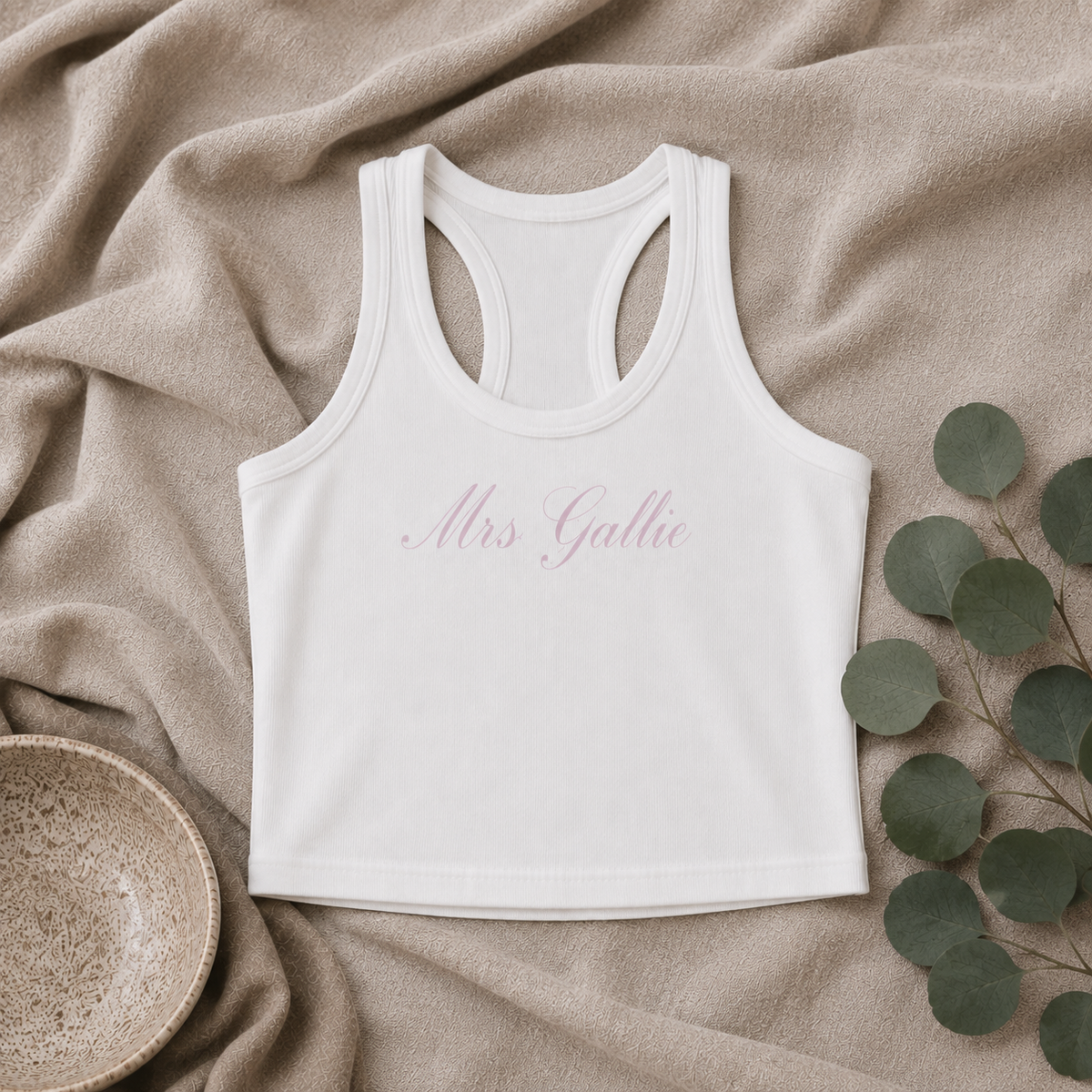 A white racerback crop top with 'Mrs. Gattie' printed in pink cursive across the chest, laid flat on a beige textured blanket with eucalyptus leaves and a ceramic bowl.
