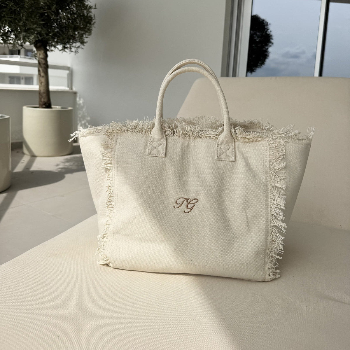 White tote bag with fringe details and a monogram on a light surface, with a neutral background.