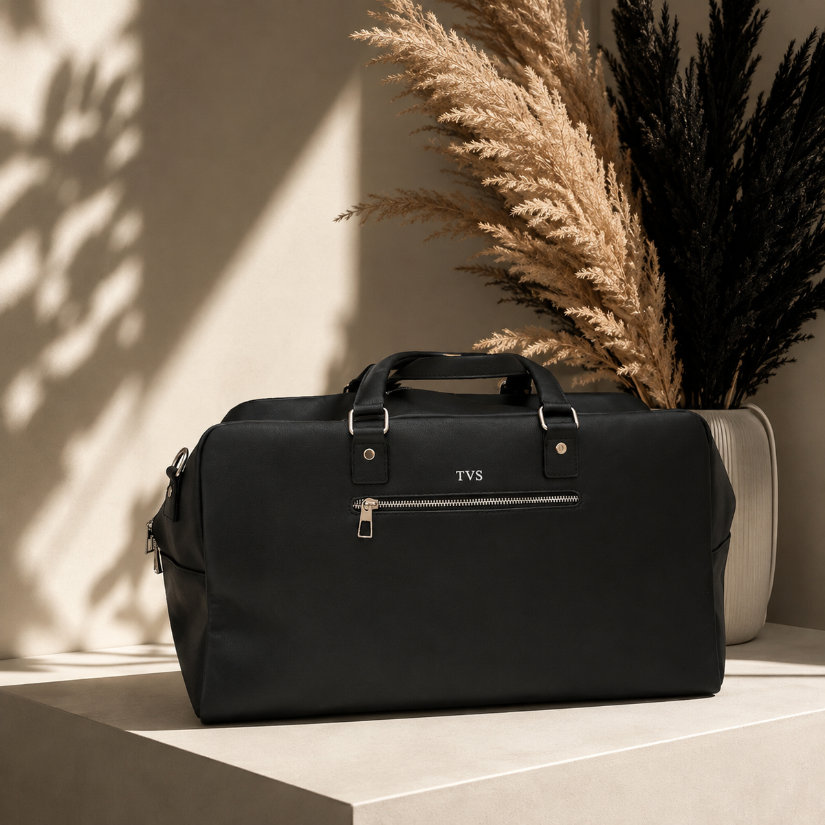 A black duffel bag with silver hardware and a front zip pocket sits on a light stone pedestal. The bag has a small silver 'TYS' logo on the front.
