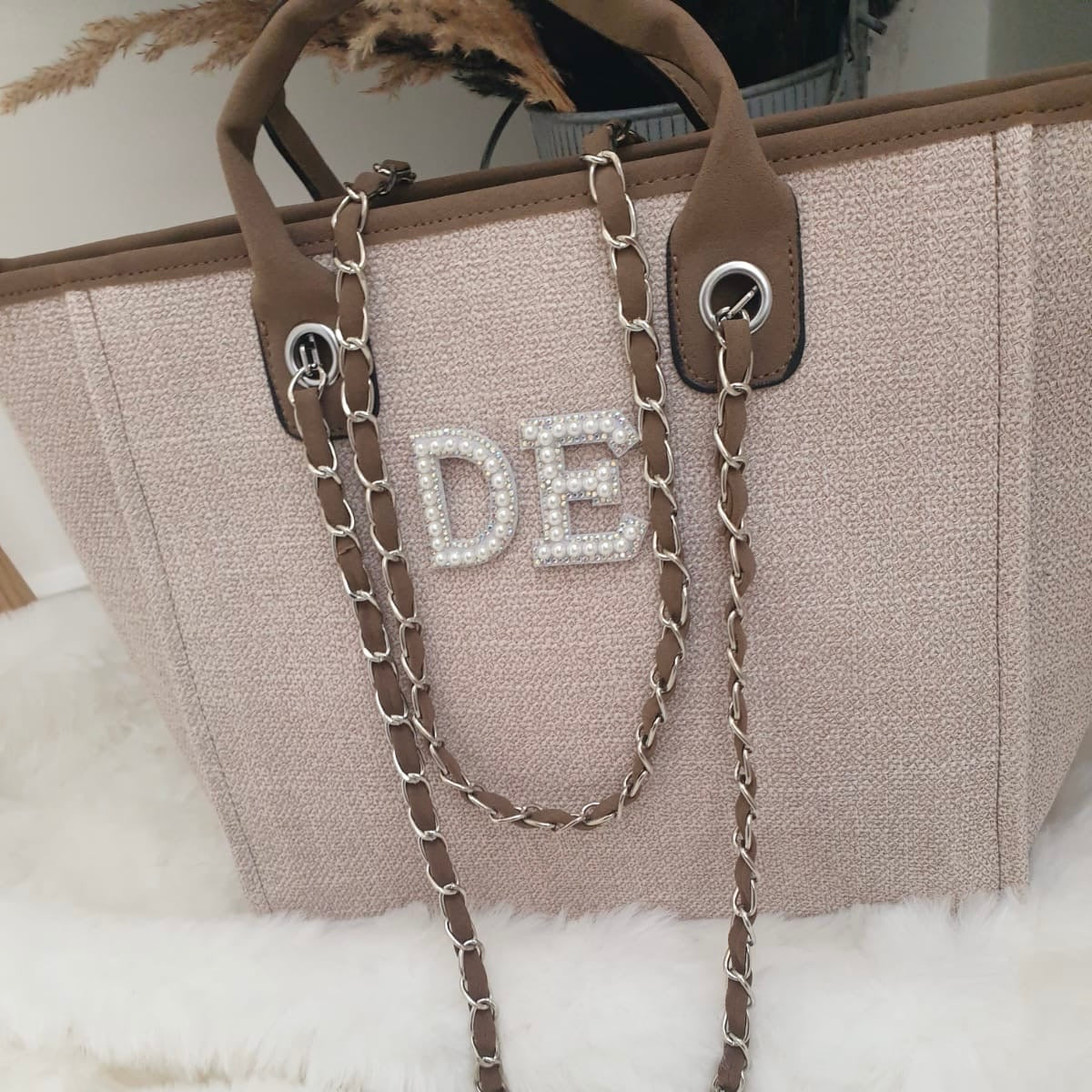 Beige handbag with decorative chain strap and 'DE' initials on a white surface.
