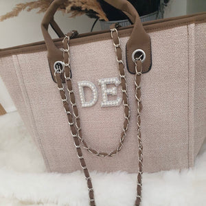 Beige handbag with decorative chain strap and 'DE' initials on a white surface.