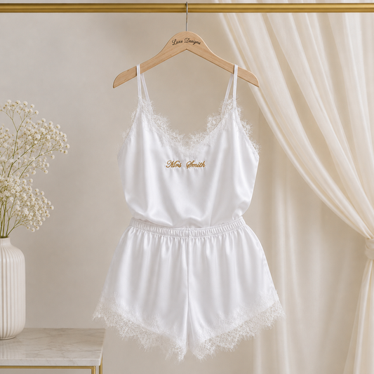 White satin lingerie set with lace trim hanging on a wooden hanger against a soft beige curtain backdrop, with a small gold embroidered script on the chest.