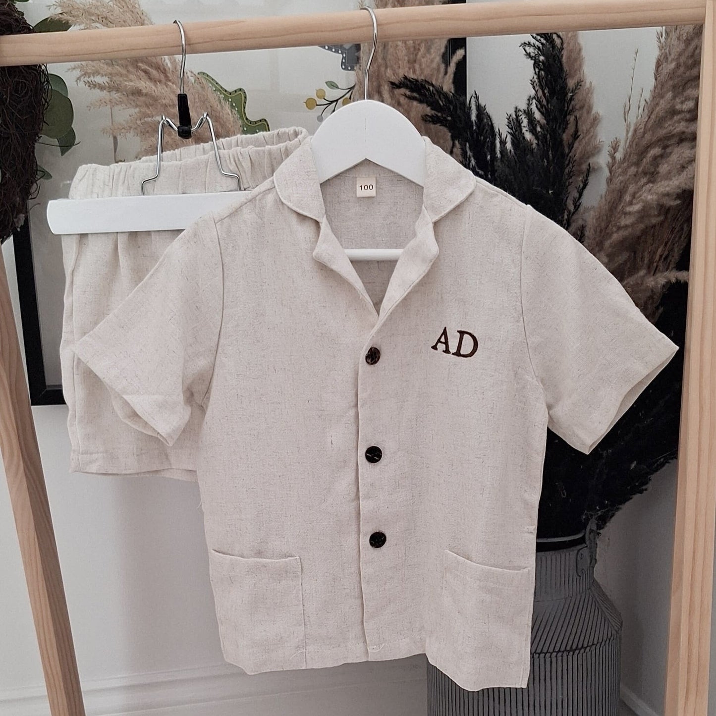Beige short-sleeve shirt with 'AD' embroidery on a hanger against a neutral background.