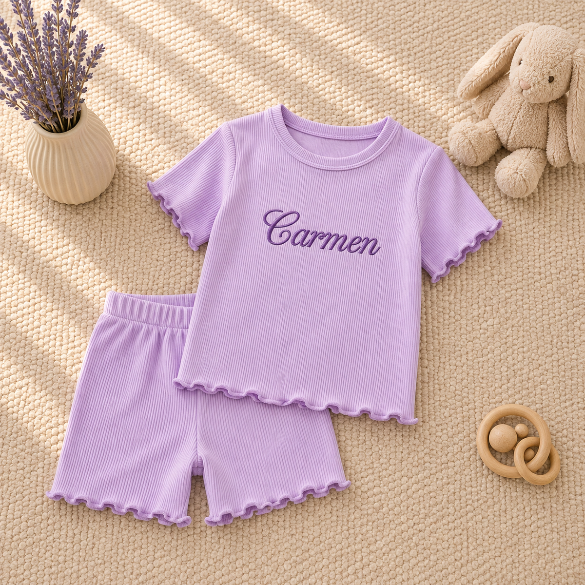 A two-piece baby outfit set in light purple, featuring a short-sleeve ribbed t-shirt with 'Carmen' embroidered on the chest and matching ribbed shorts with scalloped edges, styled on a textured beige blanket with a plush bunny, dried lavender, and a wooden teething toy.