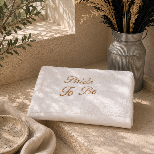 A folded white tuxedo or bridal shower towel with 'Bride To Be' embroidered in gold on the front, styled on a beige stone surface with a galvanized metal vase of dried grasses and soft natural shadows.