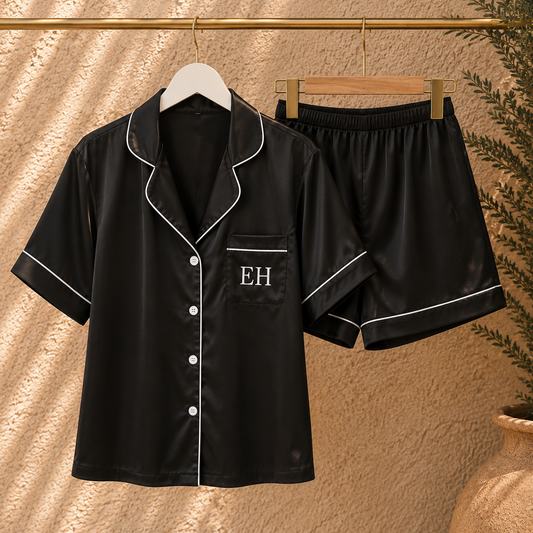 Black satin pajama set with white piping, featuring a short-sleeve button-front top with 'EH' monogram on the chest and matching shorts, displayed on hangers against a textured beige wall.
