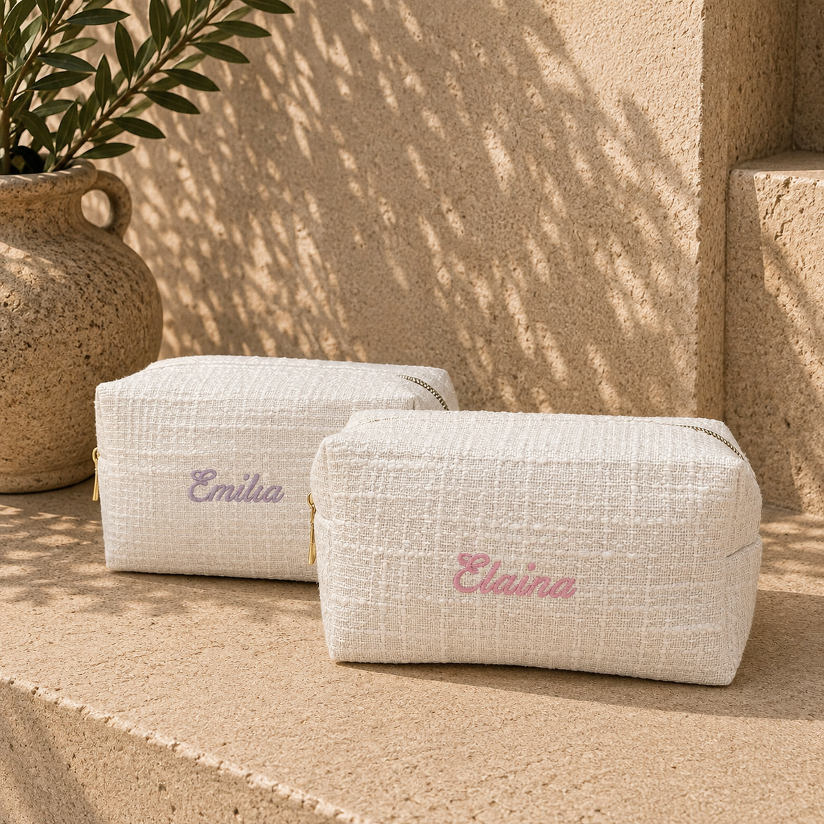 Two cream-colored woven cosmetic pouches with gold zippers, personalized with the name 'Elaina' in pink and purple embroidery, set on a stone ledge with a potted plant in the background.
