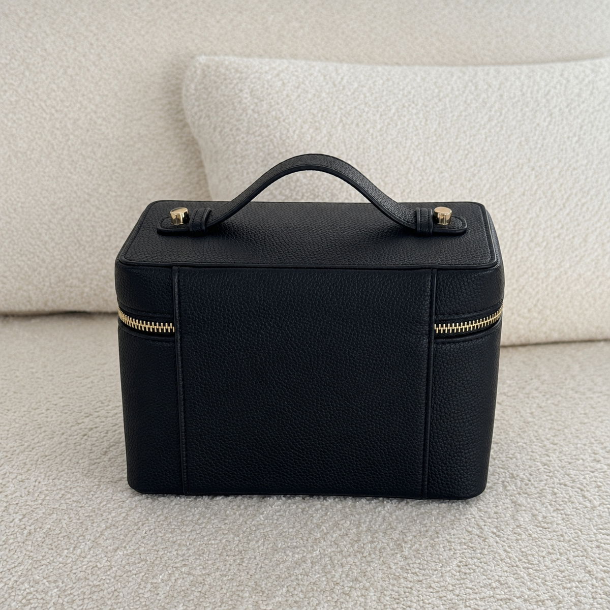 A black pebbled leather cosmetic bag with a top handle and gold-tone hardware, placed on a textured cream fabric surface.