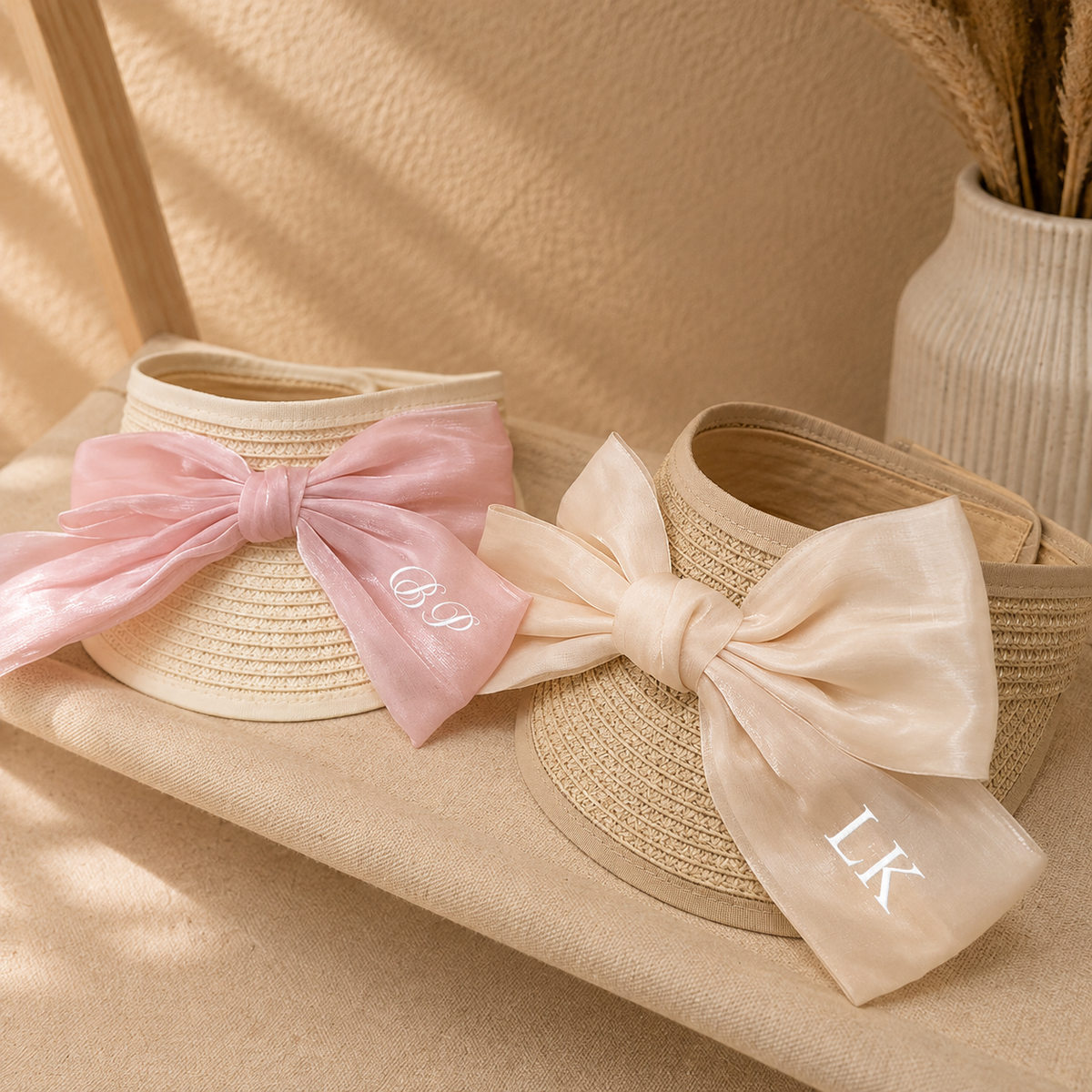 Two woven straw sun visors with large satin bows, styled on a beige fabric surface. One visor has a light pink bow with the monogram 'COP' in white, and the other has a cream-colored bow with the monogram 'LK' in white.