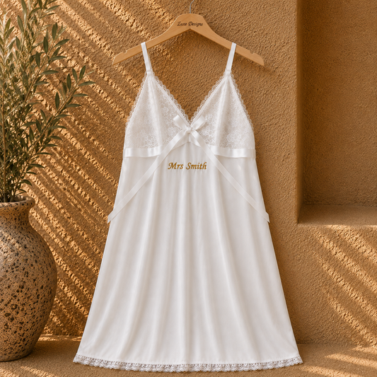 A white lace-trimmed slip dress with thin straps and a bow at the neckline, hanging on a wooden hanger against a textured stucco wall with warm sunlight and shadows. A potted plant is visible on the left.