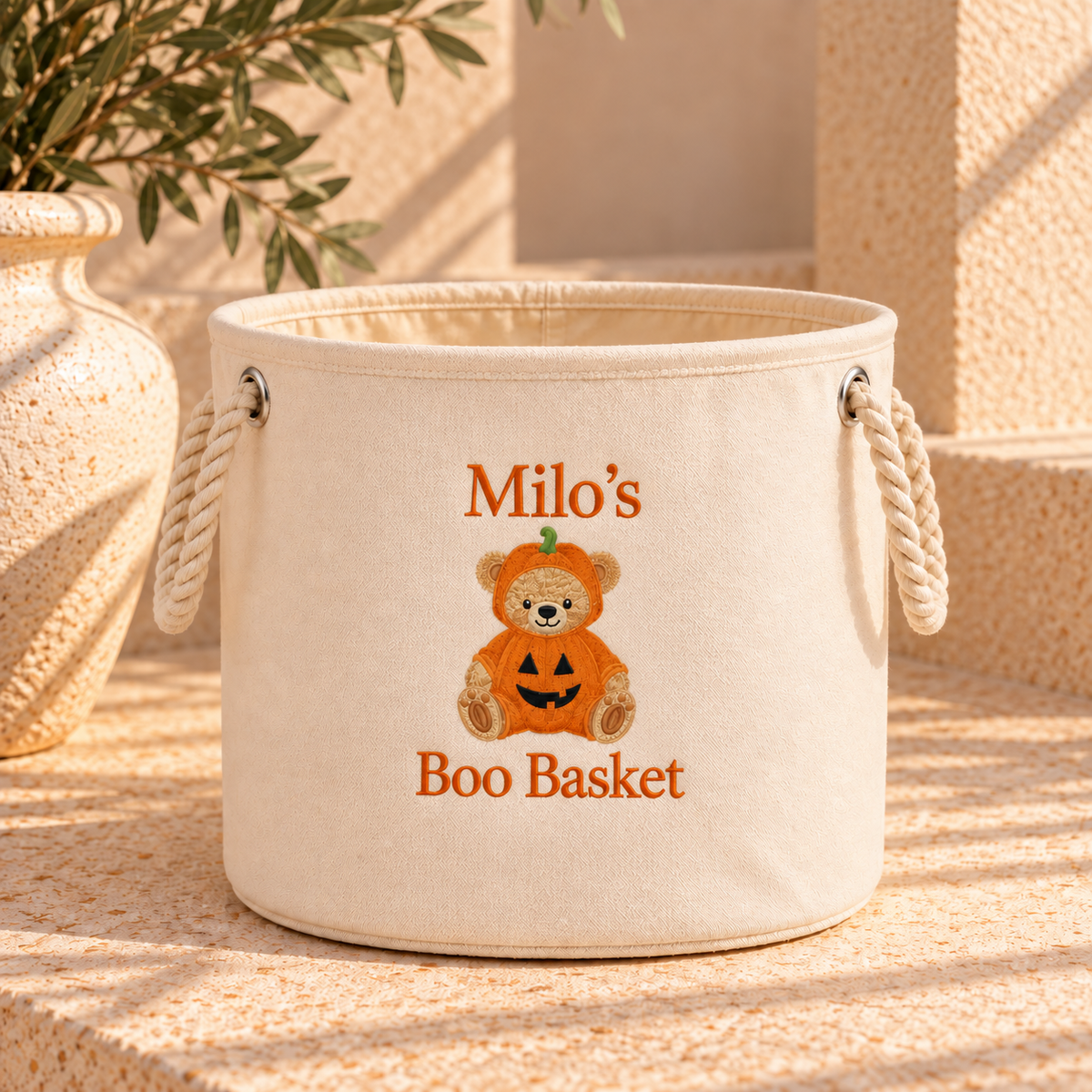 An off-white canvas storage basket with rope handles, featuring an embroidered design of a teddy bear in a pumpkin costume and the text 'Milo's Boo Basket'.