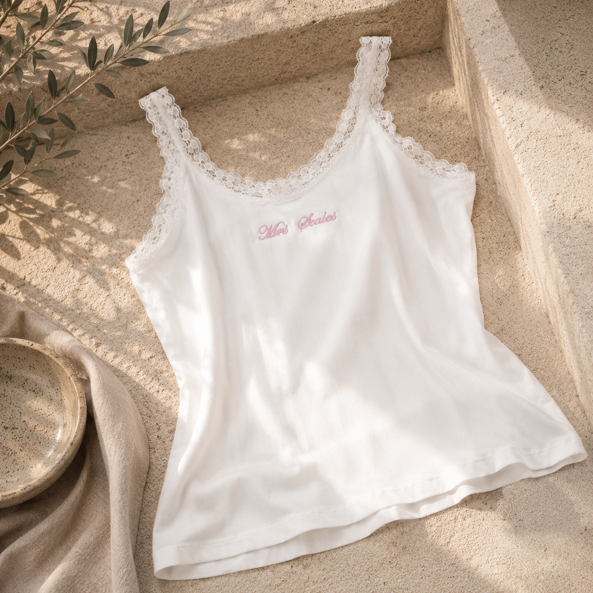 A white sleeveless camisole top with delicate lace trim along the scoop neckline and small pink script embroidery on the chest, styled on a beige stone surface with soft natural shadows.
