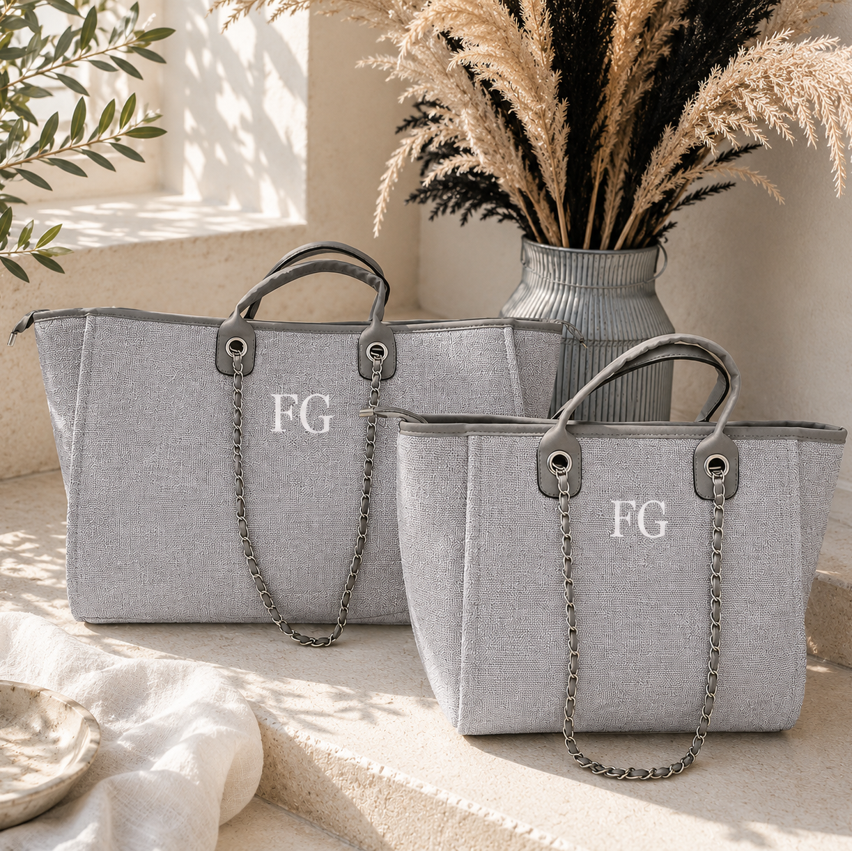 Two light gray woven tote bags with top handles, chain shoulder straps, and the letters 'FG' printed on the front, styled on a sunlit stone surface with dried grass in a vase behind them.