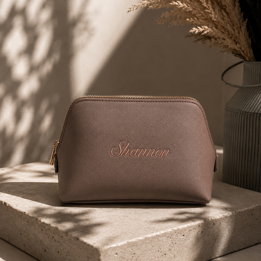 A taupe faux-leather cosmetic pouch with gold zipper trim and the name 'Shannon' embroidered in rose gold on the front, displayed on a stone slab with warm natural shadows.