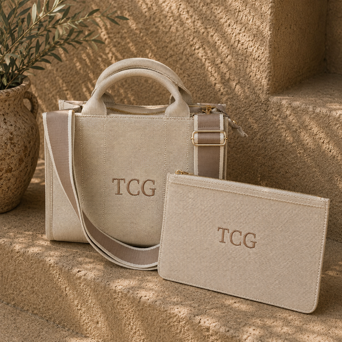 A beige textured handbag with a matching zip pouch, both embossed with the initials 'TCG', displayed on stone steps next to a potted plant.