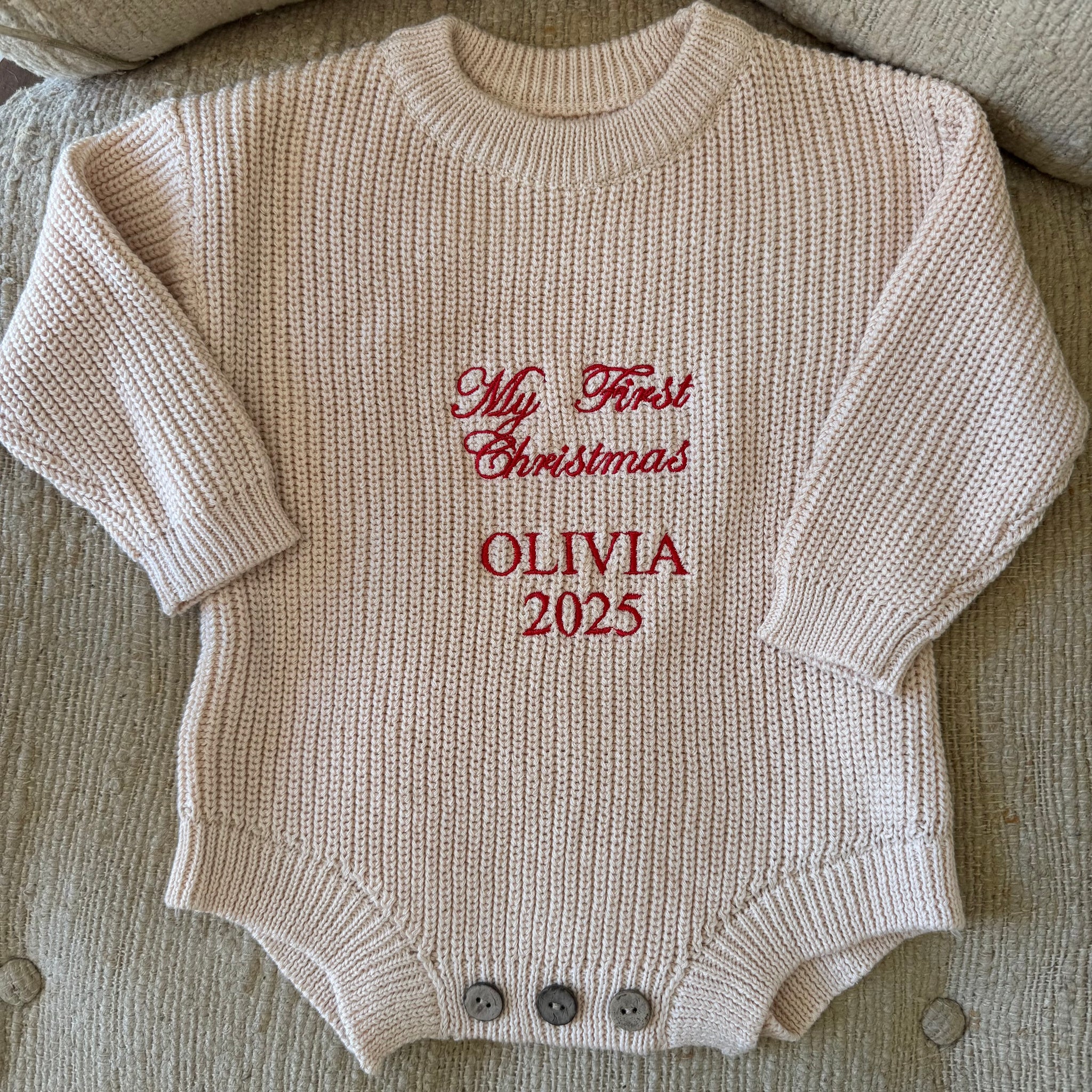 Knitted baby romper with 'My First Christmas Olivia 2025' text on a textured surface
