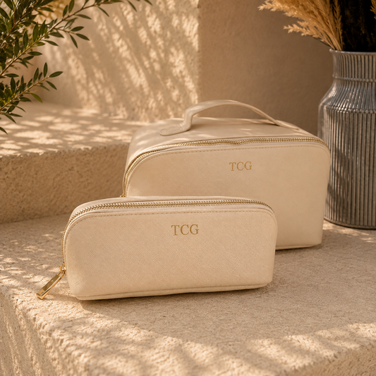 Two cream-colored zippered cosmetic bags with gold zippers and monogrammed 'TCG' logos, styled on a textured stone surface with warm natural light and shadow patterns.