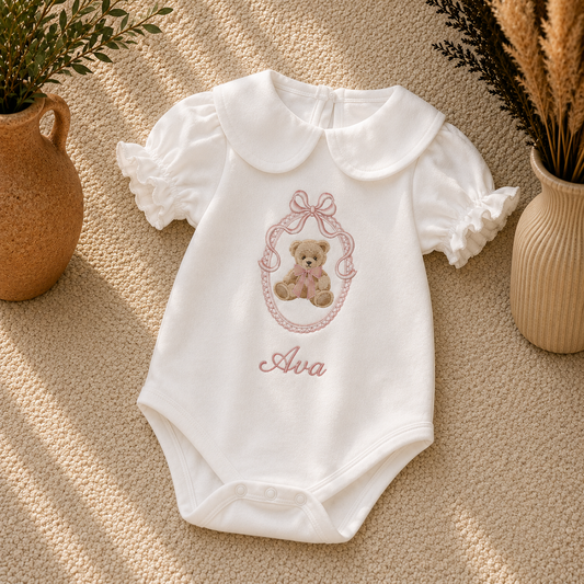 A white short-sleeve baby bodysuit with a Peter Pan collar, featuring an embroidered teddy bear in a pink bow frame and the name 'Ava' in pink script. The bodysuit is laid flat on a textured beige carpet, styled with potted plants on either side.