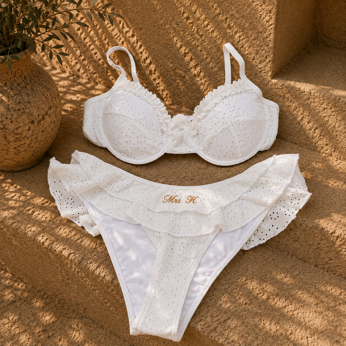 White eyelet lingerie set with ruffle trim, featuring a bra and matching thong with gold embroidered text, styled on a sandy surface with a potted plant in the background.