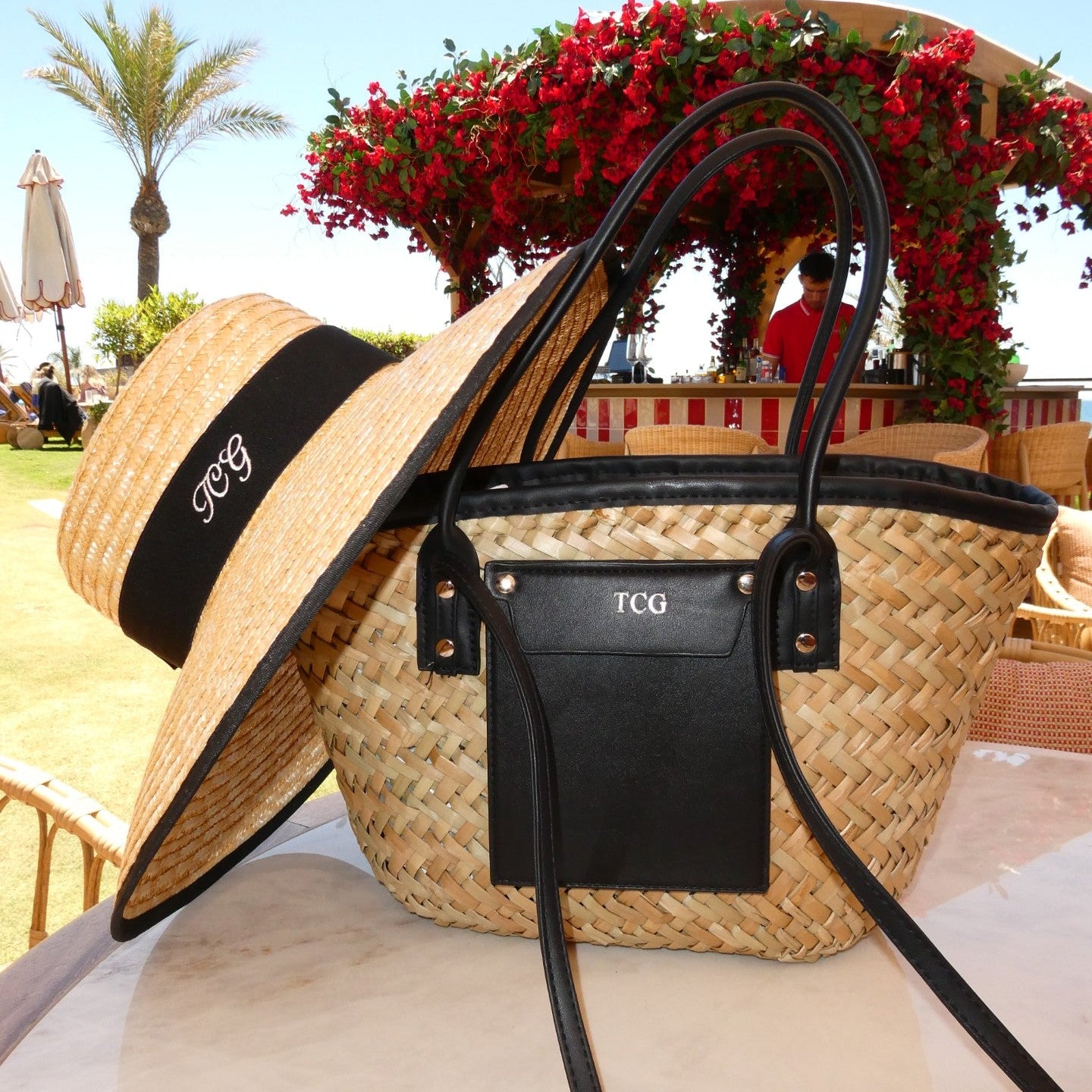 Woven straw bag with black accents and a matching hat on a beach setting.