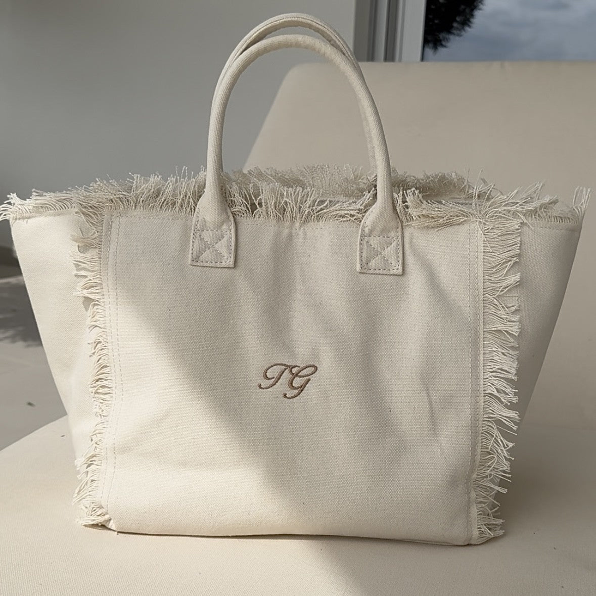 Beige tote bag with fringe details and initials on a light background
