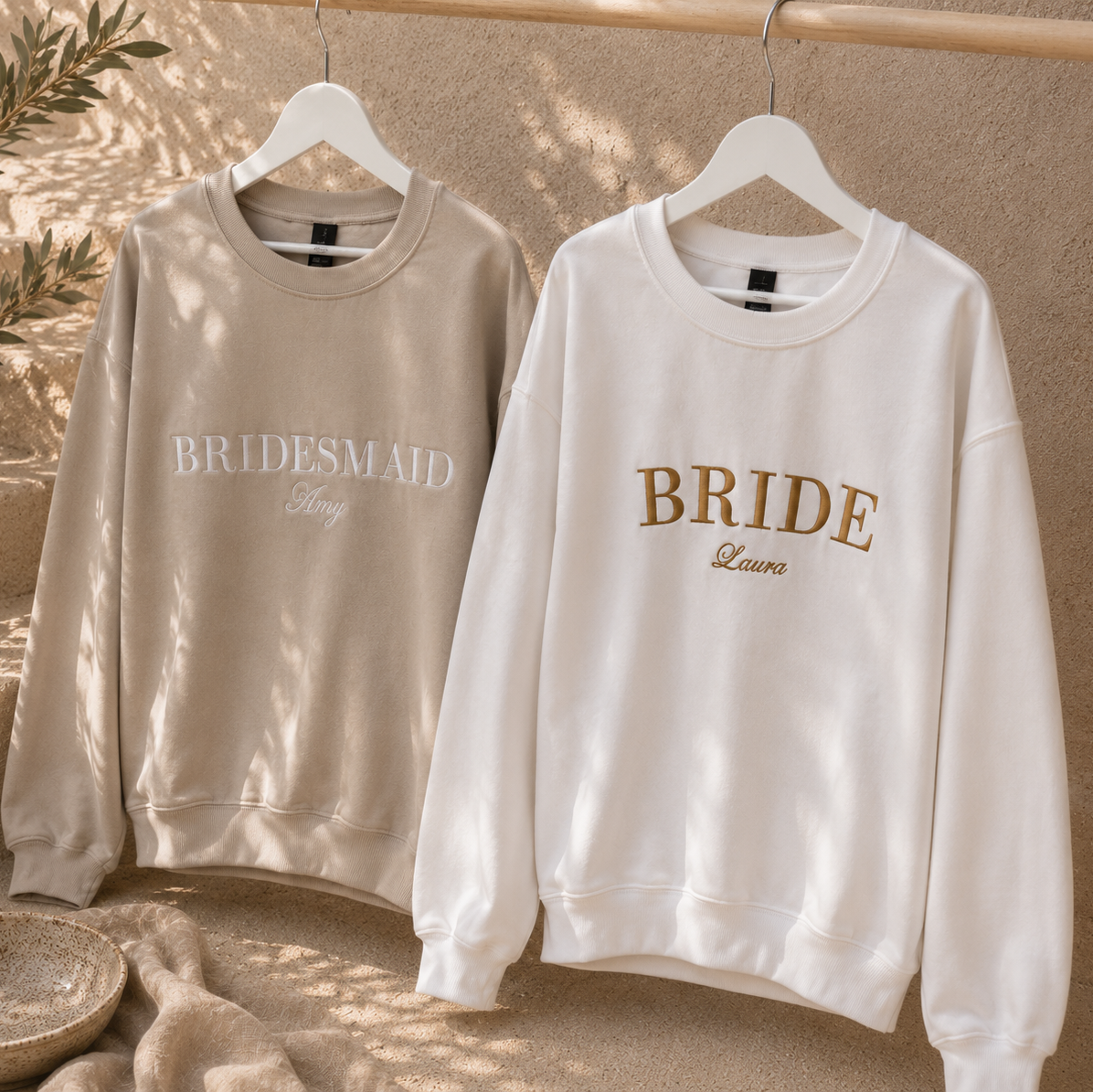 Two crewneck sweatshirts hanging on white hangers against a textured beige wall with soft leaf shadows. The left sweatshirt is light taupe with 'BRIDESMAID' in white script, and the right sweatshirt is off-white with 'BRIDE' in tan and 'Laura' in gold script.