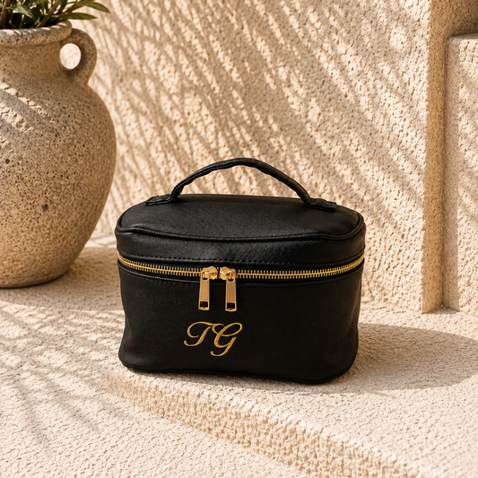 A black faux-leather cosmetic case with gold zipper pulls and a gold embroidered monogram on the front, placed on a textured beige stone surface with a ceramic pot and leaf shadows in the background.